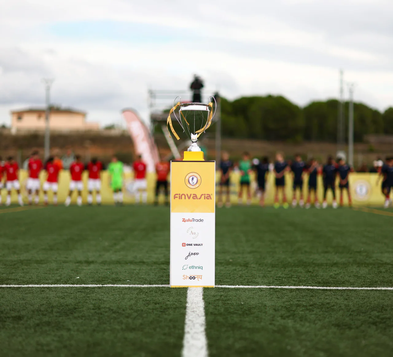 The WSG Football Cup trophy on its podium at the edge of the pitch, teams lined up behind